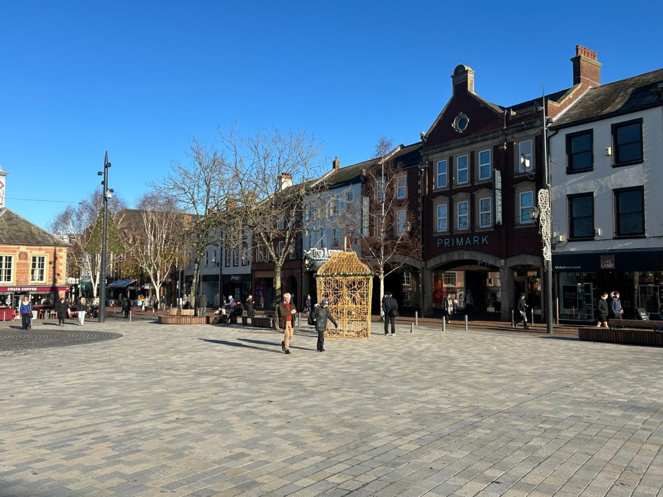 A bustling Carlisle City centre square with shops, trees, and clear blue skies.