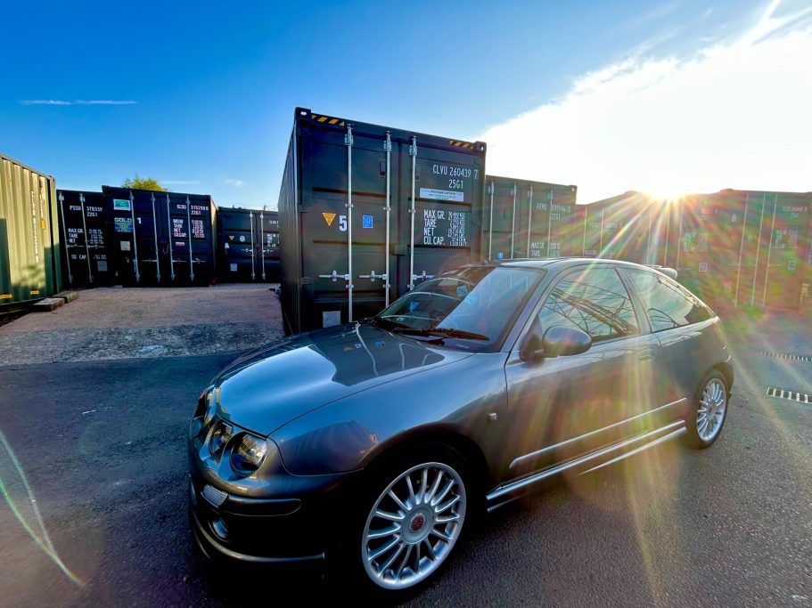 Dark hatchback car parked near shipping containers at sunset.