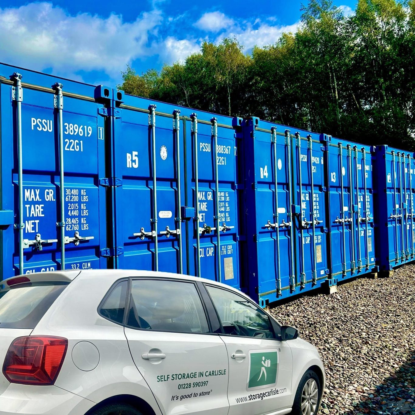Row of blue shipping containers beside a white car on gravel. Green trees in the background.