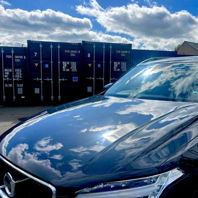Shiny black car reflecting clouds, with storage containers in the background.
