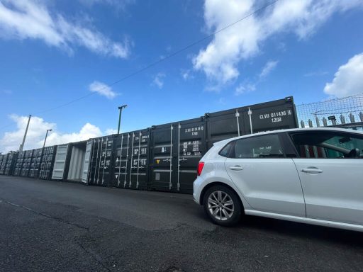 A white car parked beside a row of shipping containers under a blue sky.