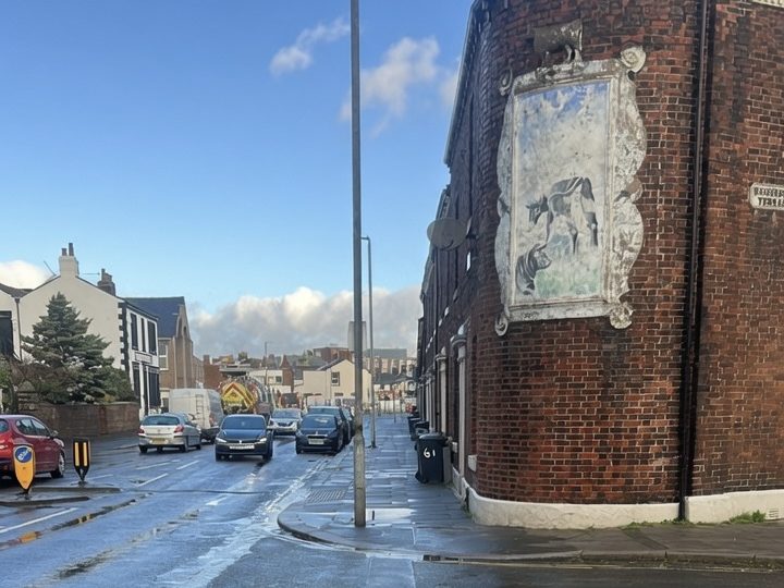 Street scene showing a brick building with a mural and parked cars under a blue sky at St. Nicholas Gate, Carlisle