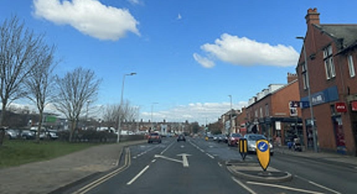 A busy road with shops and a roundabout under a clear blue sky in Carlisle.