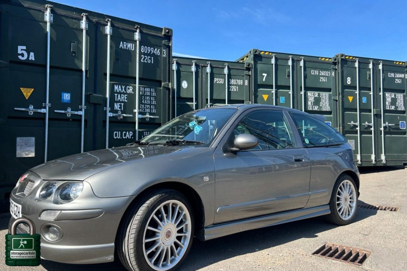 Silver hatchback car parked in front of green storage containers under a clear blue sky.