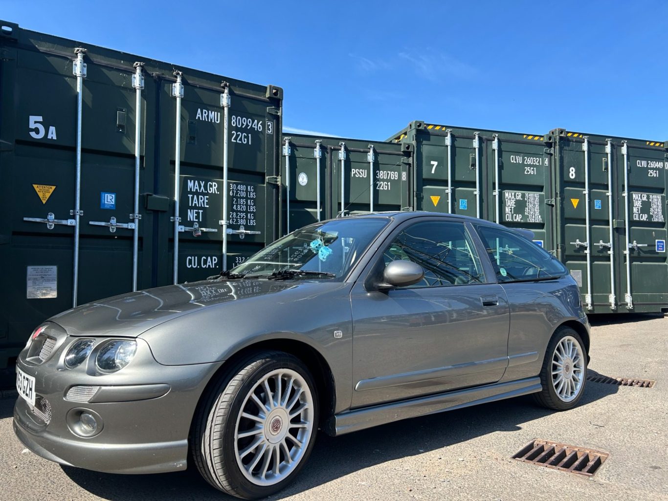 Silver hatchback car parked in front of green storage containers under a clear blue sky.